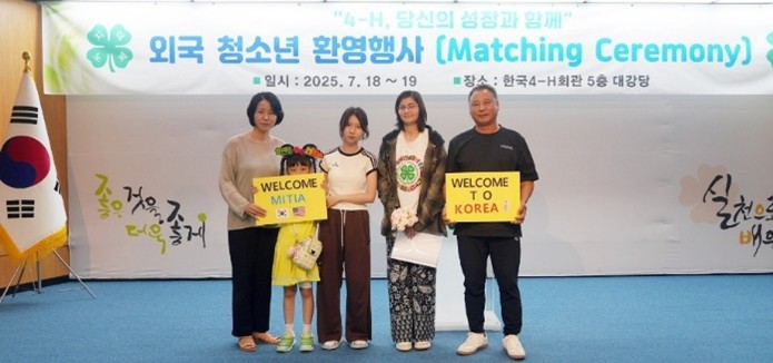 A family with signs that say Welcome to Korea and Welcome Mitia in front of a banner that reads matching ceremony.